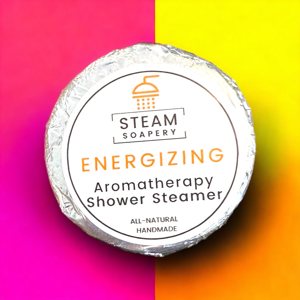 The image depicts an energizing aromatherapy shower steamer. The steamer has the word "STEAM" written on it. The steamer is circular in shape with a colorful pink, yellow, and orange background and appears to be handmade using all-natural ingredients. It promotes an energizing and refreshing experience during a shower. The steamer is designed to release natural aromatherapy scents, which can enhance relaxation and rejuvenation. This image showcases a product that provides a luxurious and invigorating shower